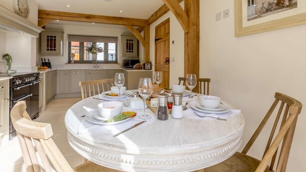 A dining area with a round table set for four, wooden chairs, a kitchen in the background, and a framed picture on the wall. - 2 Bed in Newton Tracey (oc-80381) (Newton Tracey)
