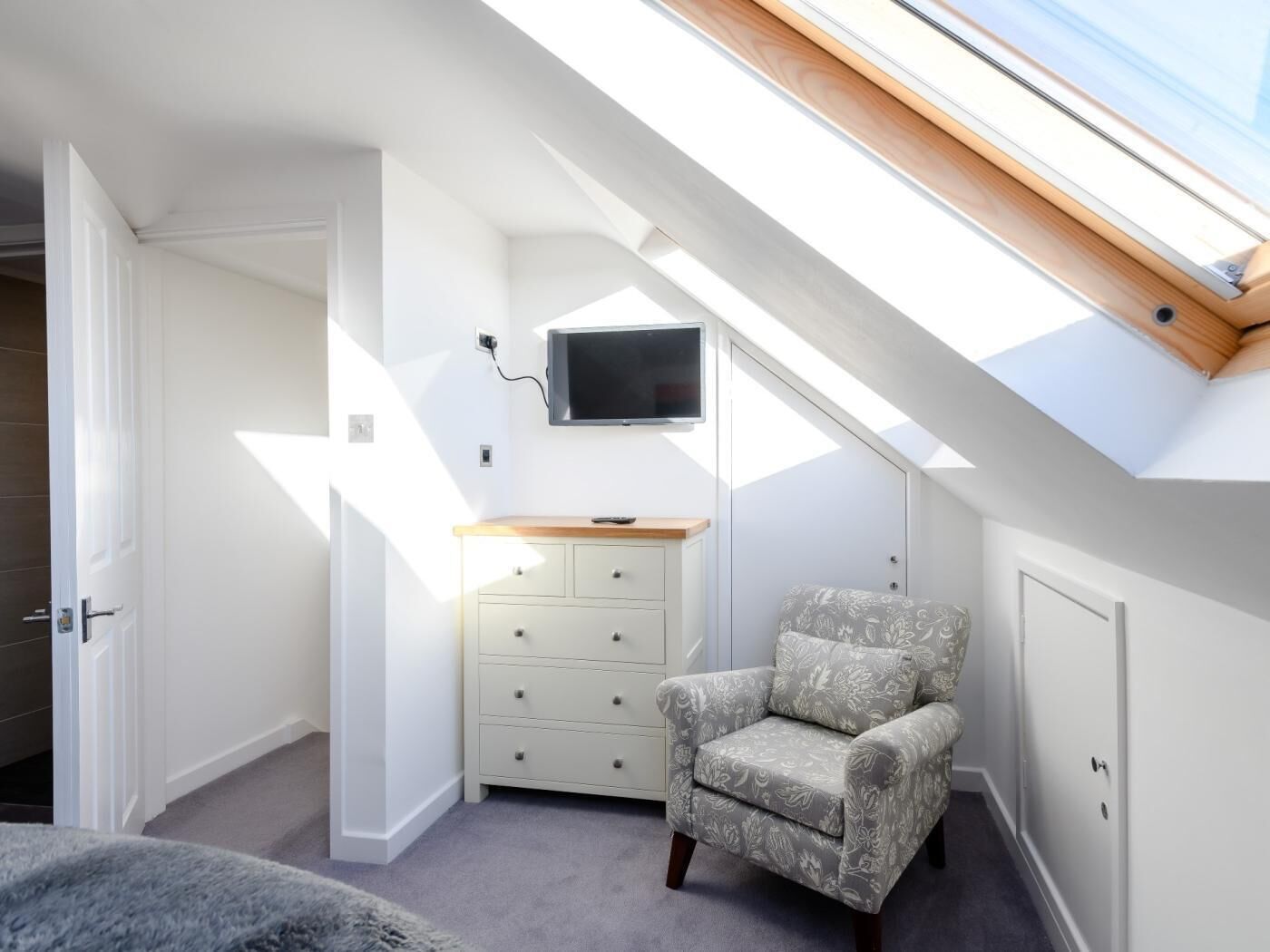 A room with a sloped ceiling, a television mounted on the wall, a wooden cabinet, a patterned armchair, and a door.
