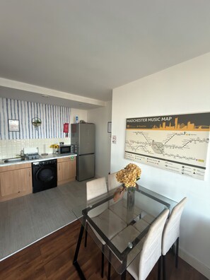 A modern kitchen with a dishwasher, refrigerator, and a dining area with a glass table and chairs. - Manchester City Centre/ Prime Location Apartment (Salford)