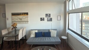 Een moderne woonkamer met een glazen salontafel, een bank met kussens, een eettafel met stoelen en een stadsgezicht door grote ramen.