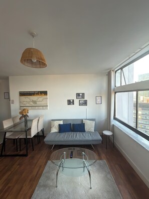 A modern living room with a glass coffee table, a sofa with cushions, a dining table with chairs, and a cityscape view through large windows. - Manchester City Centre/ Prime Location Apartment (Salford)