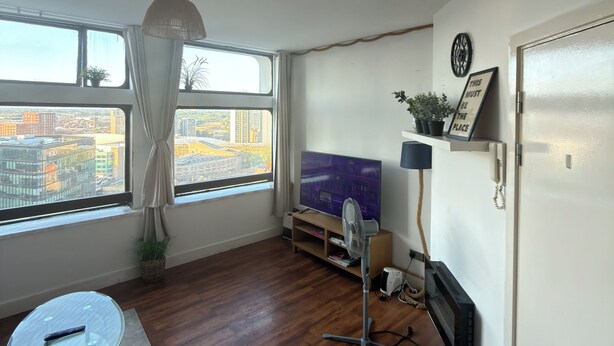 Een kamer met een groot raam, een flatscreen televisie, een ventilatorkachel, een kleine glazen tafel en een deur.