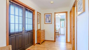 A hallway with wooden floors, a dark door with blinds, a wooden dresser, and a dining area in the background.