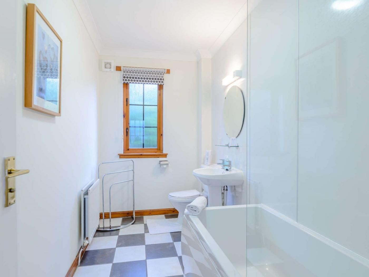 Un baño con piso a cuadros, inodoro, lavamanos, ducha y ventana con cortina estampada.