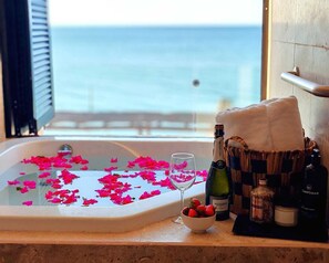 Una bañera llena de agua y pétalos de rosa, una copa de vino y distintos artículos de aseo en una encimera con vistas al mar.