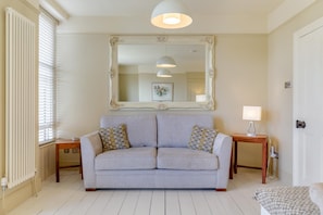 A room with a light grey sofa, two patterned cushions, a large ornate mirror, a small table with a lamp, and a radiator. - 2 Bed in Brixham (oc-80453) (Brixham)