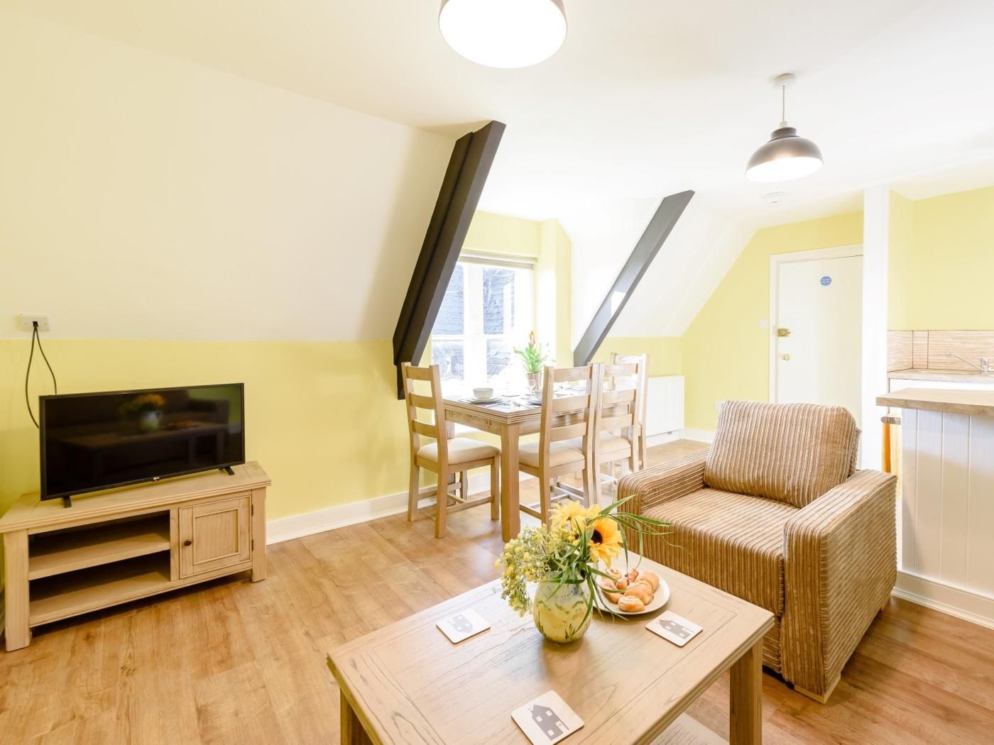 A living room with a television, dining table, chairs, a sofa, and a kitchen area with a sink.