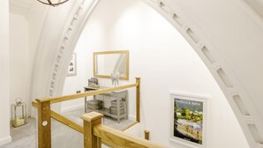 Um quarto com pé-direito alto, uma escada de madeira, um candelabro, um espelho e um cartaz emoldurado.