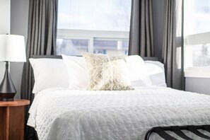 Un lit bien fait, recouvert d’une couette blanche, avec un oreiller à motifs et une lampe de chevet posée sur une table de nuit en bois.