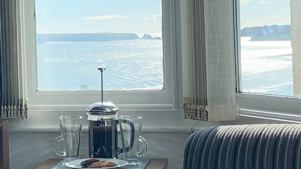 A room with a large window overlooking a snowy landscape, a coffee maker, two glasses, and a plate with cookies. - 2 Bed in Tenby (oc-fb036) (Tenby)