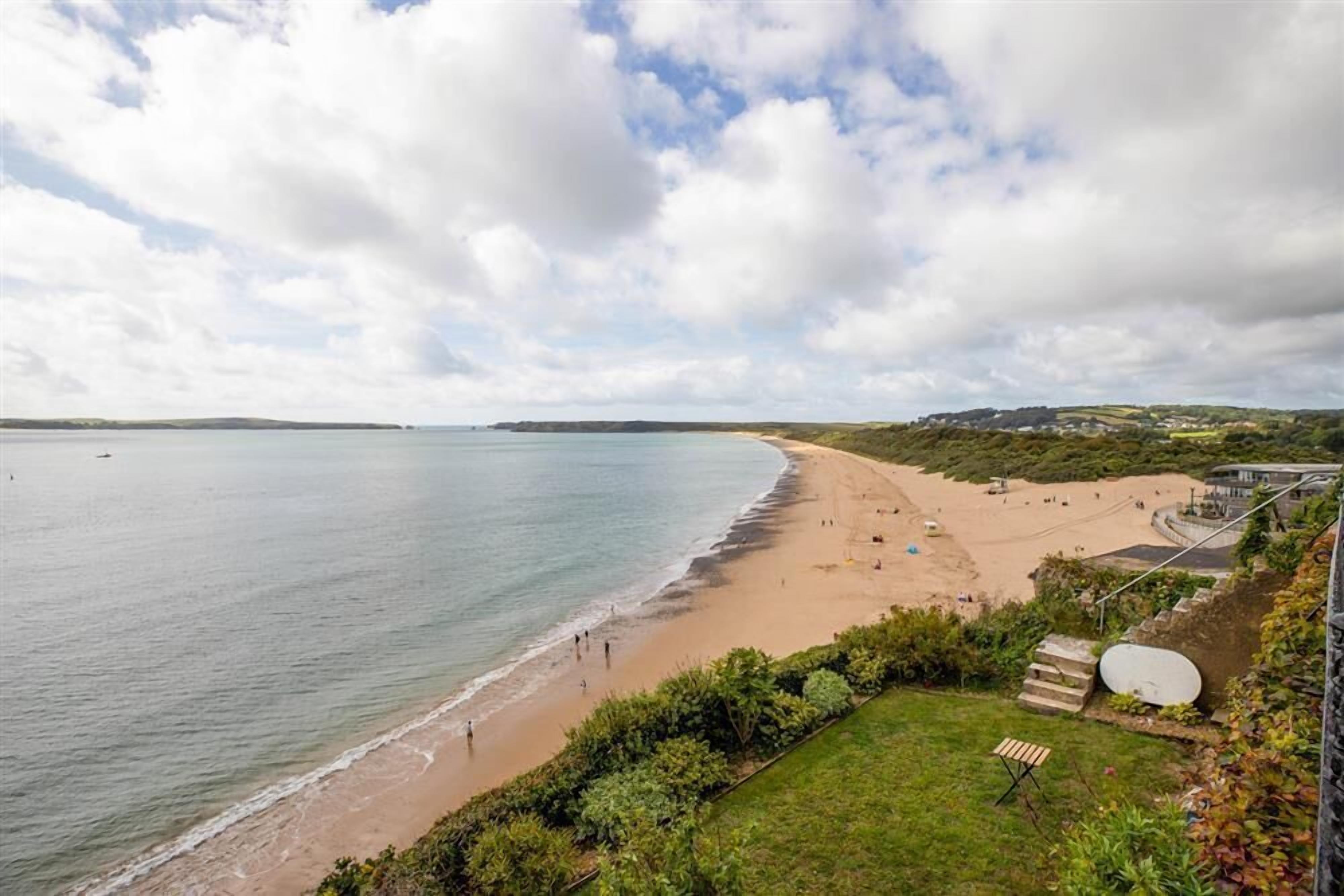 A coastal beach with a clear view of the sea, a sandy shore, and a grassy area with steps leading down to the beach.