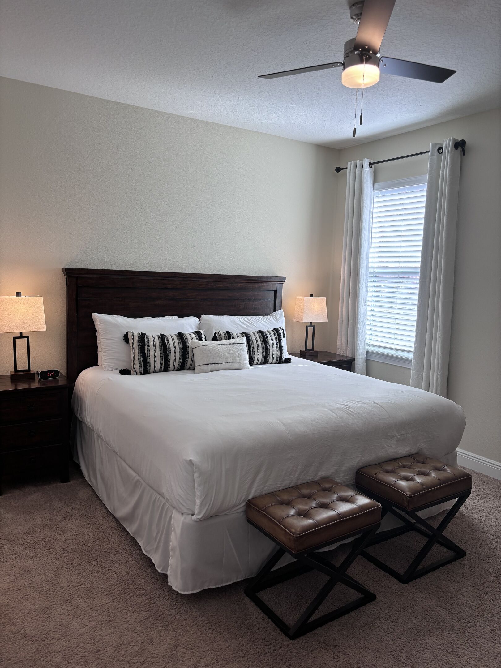 A bedroom with a large bed, two bedside tables, a ceiling fan, and a window with blinds.