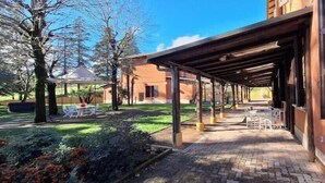 A covered outdoor seating area with tables and chairs, a garden with shrubs and a small tree, and a building with a brick facade. - Corina's Place Bologna Villa (Bologna)