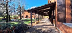 Un área de estar al aire libre con techo, mesas y sillas, un jardín con arbustos y un árbol pequeño, y un edificio con fachada de ladrillo.