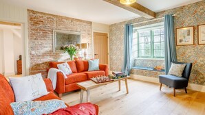 Ein Wohnzimmer mit Backsteinwand, Holzbalkendecke, einer Couch, einem Sessel, einem Couchtisch und einem Fenster mit Vorhängen.