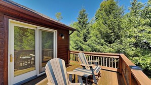 En træterrasse med Adirondack-stole og et bord, en skydedør samt udsigt til træer.