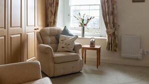 A room with a beige armchair, a small wooden table, a radiator, and a window with curtains. - 1 Bed in Tenby (oc-fb024) (Tenby)