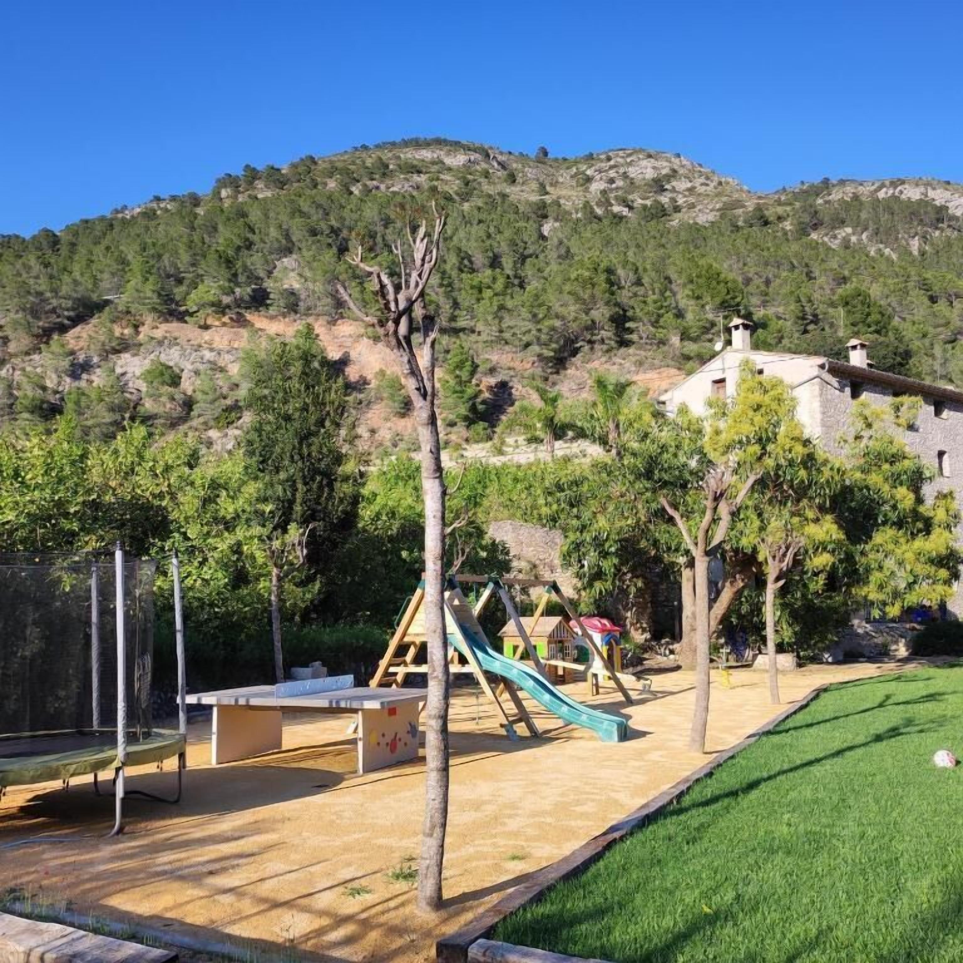 Une aire de jeux avec des structures d’escalade et un toboggan, entourée d’arbres et d’une pelouse, avec une montagne en arrière-plan.