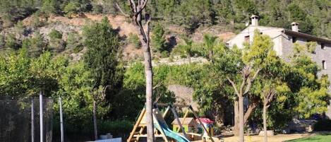 Une aire de jeux avec des structures d’escalade et un toboggan, entourée d’arbres et d’une pelouse, avec une montagne en arrière-plan.
