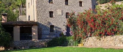 A stone building with a flat roof, a small balcony, and a garden with red flowers.