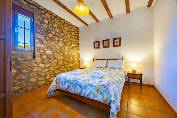 A bedroom with a stone wall, wooden beams, a bed with a floral bedspread, two bedside lamps, and a window with a cross.