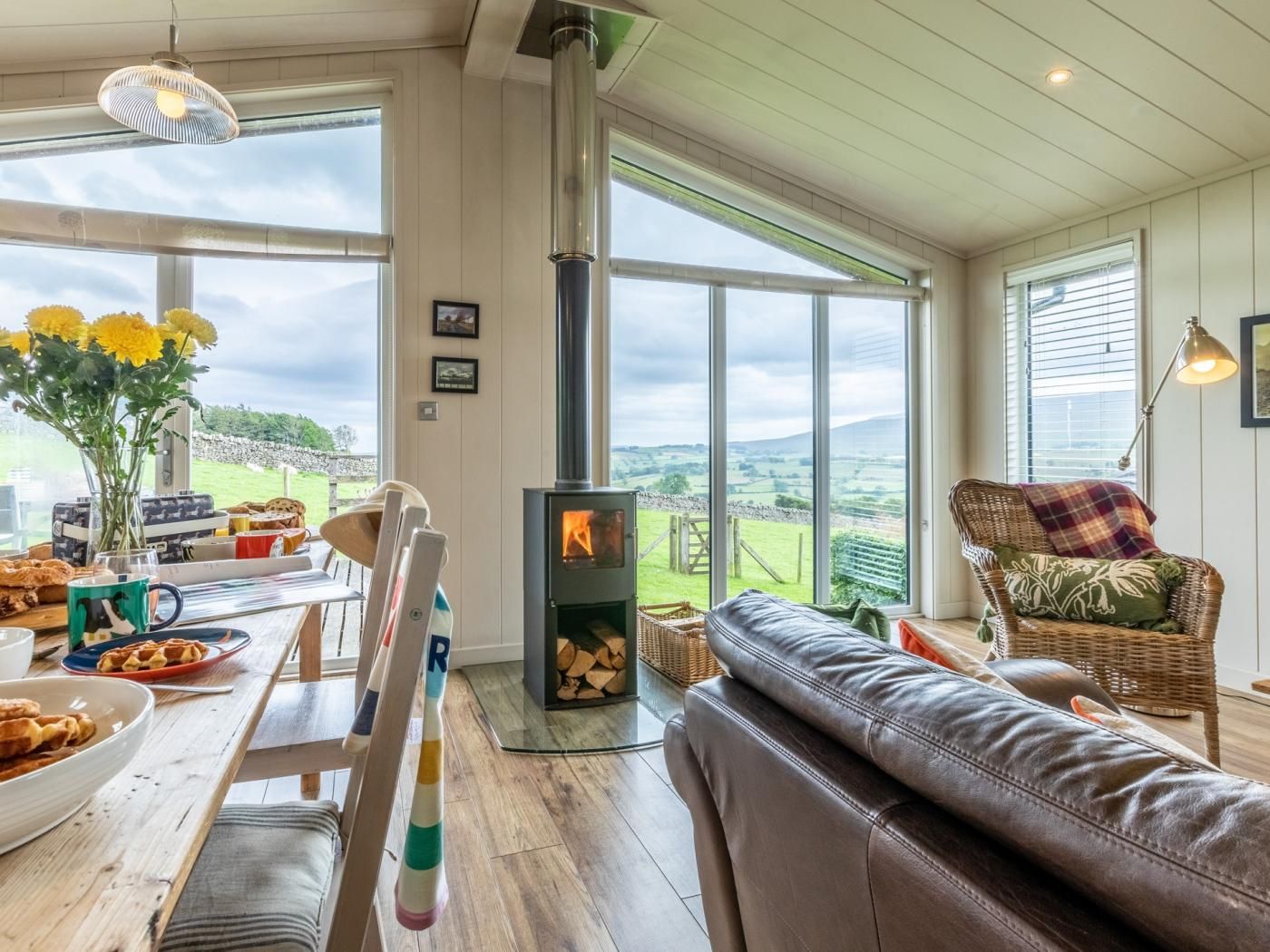 A cozy living room with a fireplace, a sofa, a dining table with breakfast items, and large windows offering a view of the countryside.