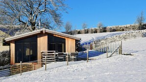 A wooden cabin with a metal roof, surrounded by snow-covered terrain and a stone wall. - 2 Bed in Wigton (oc-80563) (Caldbeck)