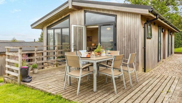 A wooden cabin with a deck, a table with chairs, and a large window. - 2 Bed in Wigton (oc-80563) (Caldbeck)
