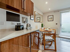 A modern kitchen with wooden cabinets, a countertop, a toaster, and a dining area with a table and chairs. - 1 Bed in Tenby (oc-fb052) (Tenby)