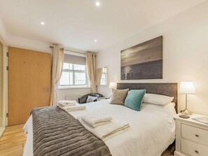 A hotel room with a bed, bedside lamps, a window with curtains, a painting on the wall, and a wooden door. - 3 Bed in Tenby (oc-fb055) (Tenby)