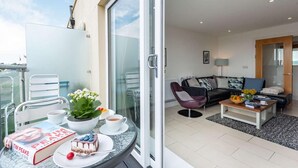 A modern living room with a sofa, coffee table, and a balcony with a table set for tea and a book. - 3 Bed in Tenby (oc-fb055) (Tenby)