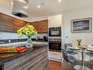 A modern kitchen with wooden cabinets, a built-in oven, a dining area with a glass table and chairs, and a vase of flowers on the counter. - 2 Bed in Tenby (oc-fb108) (Tenby)