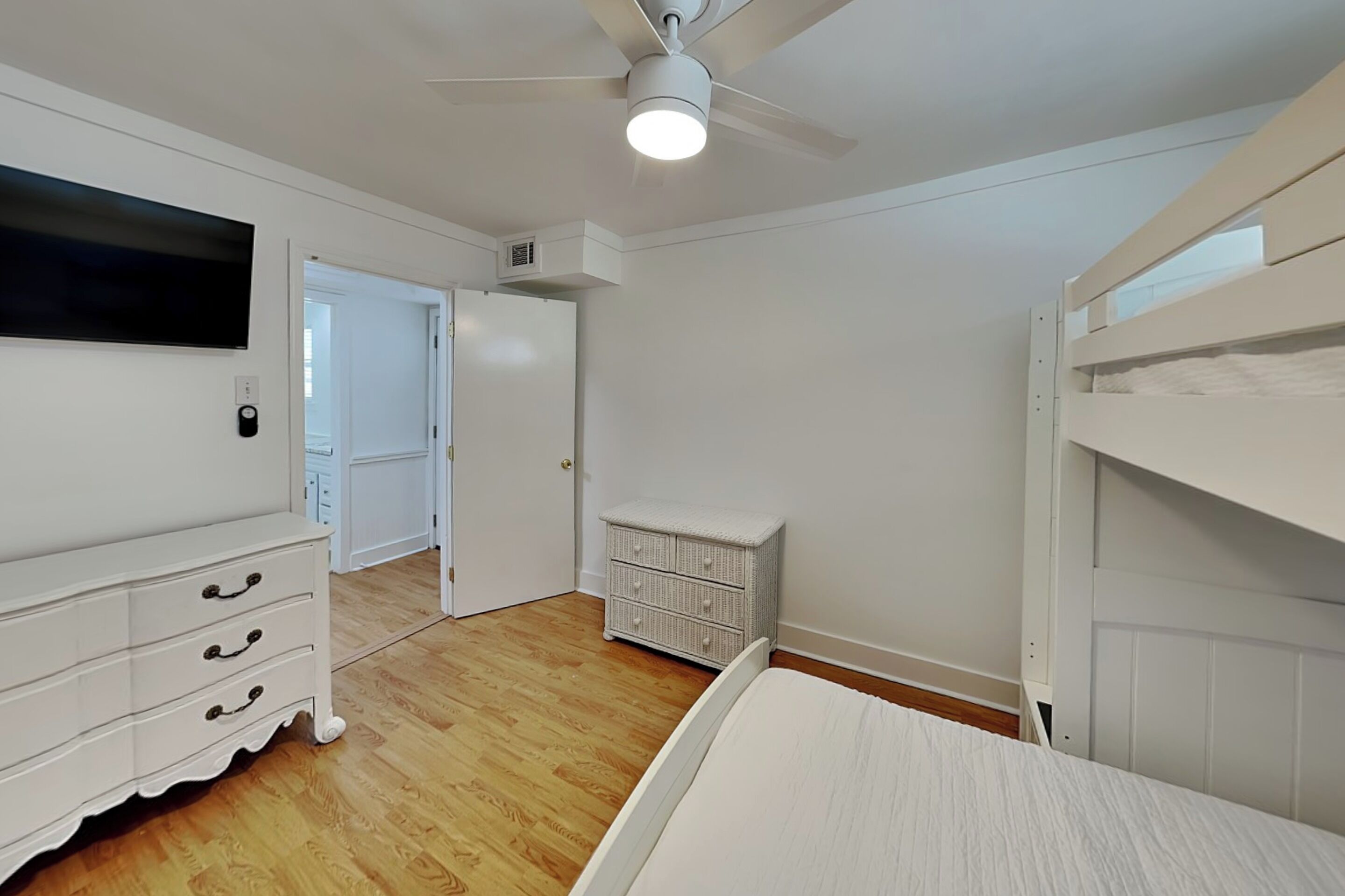 Ein Schlafzimmer mit einem Bett, einem Schminktisch, einem Fernseher, einem Deckenventilator und einer Tür zu einem anderen Raum.