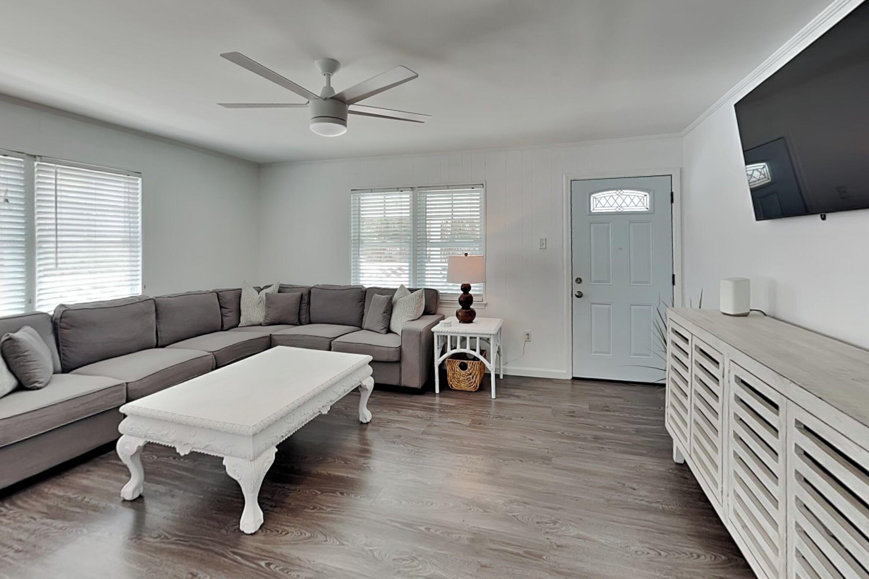 A living room with a sectional sofa, a white coffee table, a ceiling fan, a television mounted on the wall, and a door with a decorative glass panel.