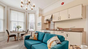 A modern living room with a teal sofa, a dining area with a table, and a kitchen with cabinets and a microwave. - 1 Bed in Tenby (oc-fb154) (Tenby)