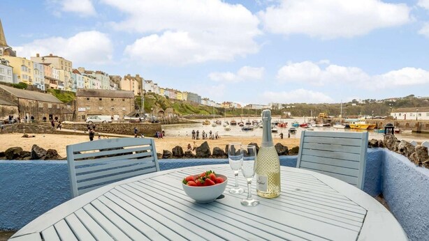 Une table installée au bord de la mer, avec une bouteille, des verres et un saladier rempli de fruits.