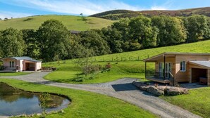 A rural property with a gravel driveway, a small pond, and two wooden buildings with attached porches. - 1 Bed in Llangammarch Wells (oc-bn089) (Treflys)