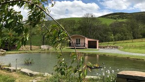 A lakeside cabin with a porch, a small dock, and a grassy area with trees. - 1 Bed in Llangammarch Wells (oc-bn089) (Treflys)