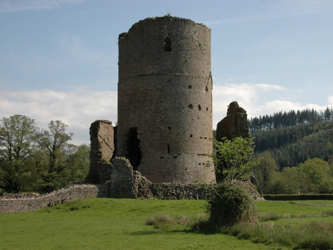 A historic stone tower with a circular base and a narrow top, surrounded by a grassy field and trees.
