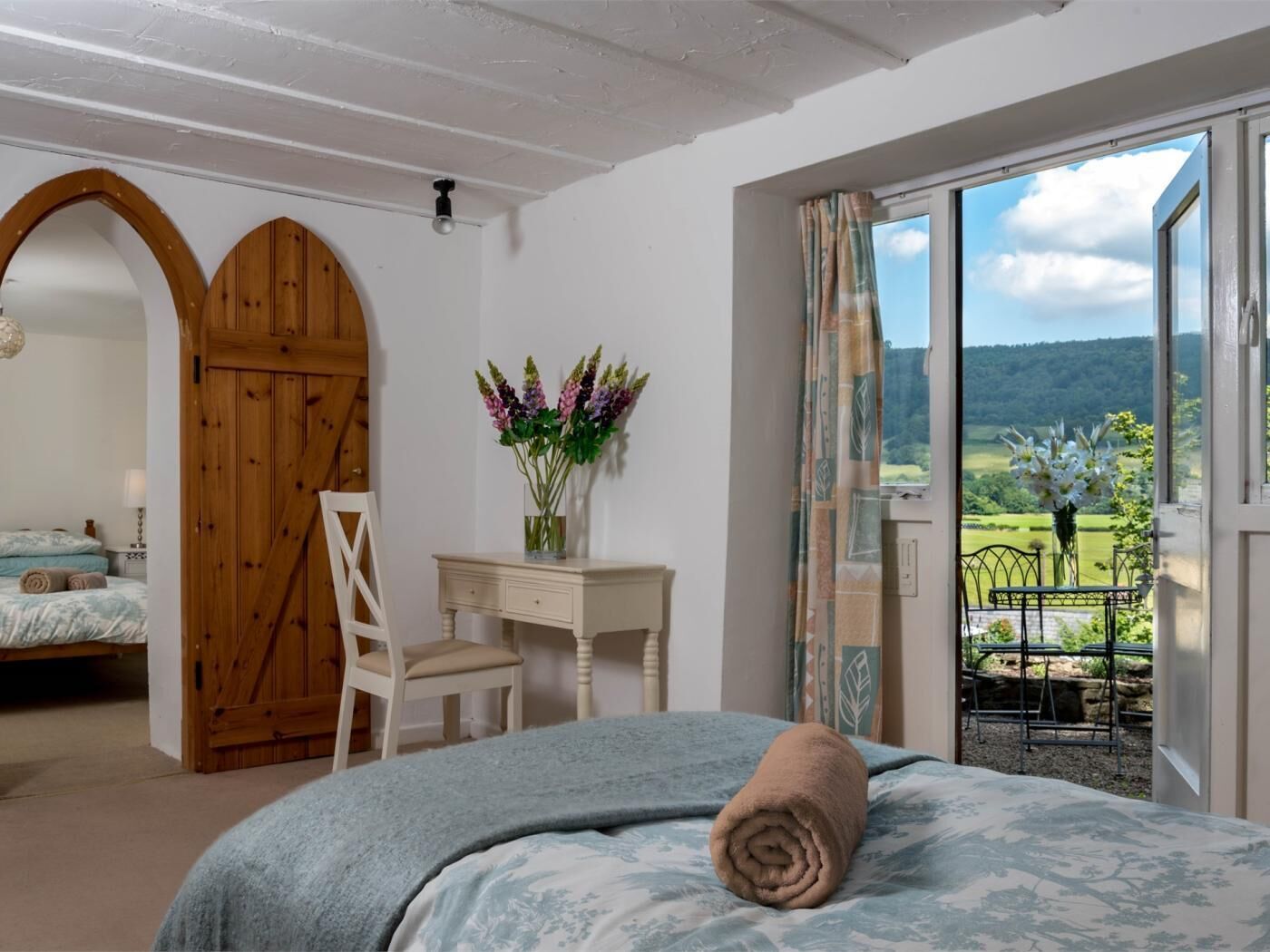 A bedroom with a bed, a desk, a chair, and a view of a green landscape through a window.
