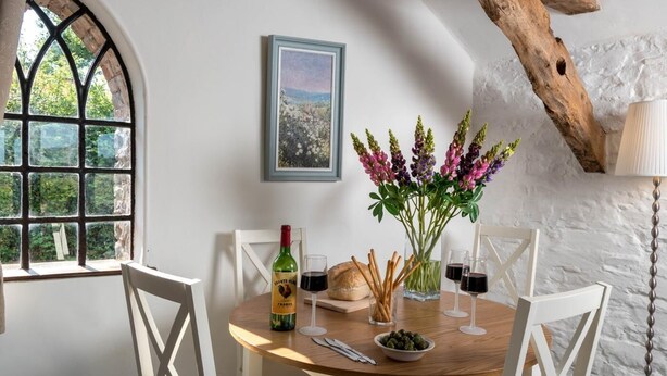 A dining area with a round table, white chairs, a bottle of wine, glasses, bread, and a vase with flowers. - 2 Bed in Brecon (oc-bn102) (Talybont-on-Usk)