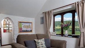 A cozy living room with a brown sofa, a map on the wall, a window with a view of greenery, and a vase with flowers on the windowsill. - 2 Bed in Brecon (oc-bn102) (Talybont-on-Usk)