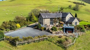 Une maison en pierre avec une allée pavée, une cour clôturée et une vue sur une colline verdoyante où paissent des moutons.