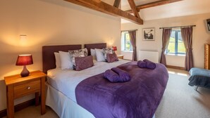 Une chambre avec un grand lit, des poutres apparentes, une table de chevet avec une lampe, une fenêtre avec des rideaux et un radiateur.