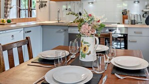 Ein gedeckter Esstisch mit Tellern, Gläsern und Besteck, eine Vase mit Blumen und eine Küche im Hintergrund.