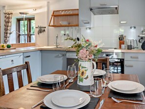 A dining table set with plates, glasses, and cutlery, a vase with flowers, and a kitchen in the background. - 2 Bed in Crickhowell (oc-bn194) (Cwmdu)