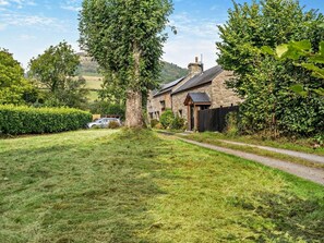 A stone house with a paved path leading to it, surrounded by greenery and a car parked in the yard. - 2 Bed in Crickhowell (oc-bn194) (Cwmdu)