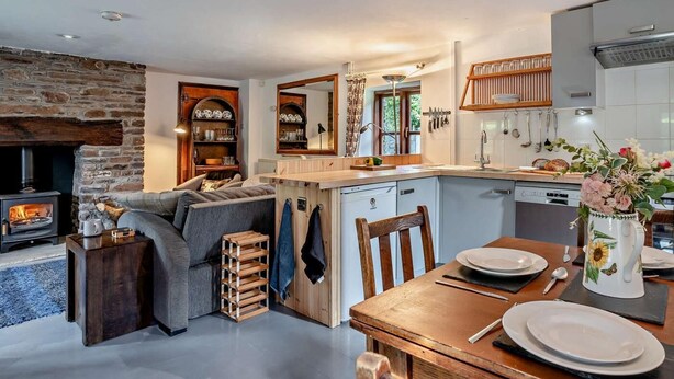 Ein gemütliches Wohnzimmer mit Kamin, eine Essecke mit Holztisch und Stühlen sowie eine Küche mit weißen Schränken und einer Steinwand.