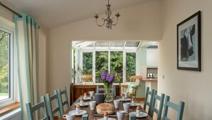 Comedor con mesa de madera dispuesta para el desayuno, sillas, una lámpara de araña y vistas a través de un invernadero.