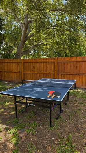 An outdoor ping pong table with two paddles and a ball, surrounded by a wooden fence and trees. - Flamingo House, Cozy 2/2 [pet friendly] (Sarasota)